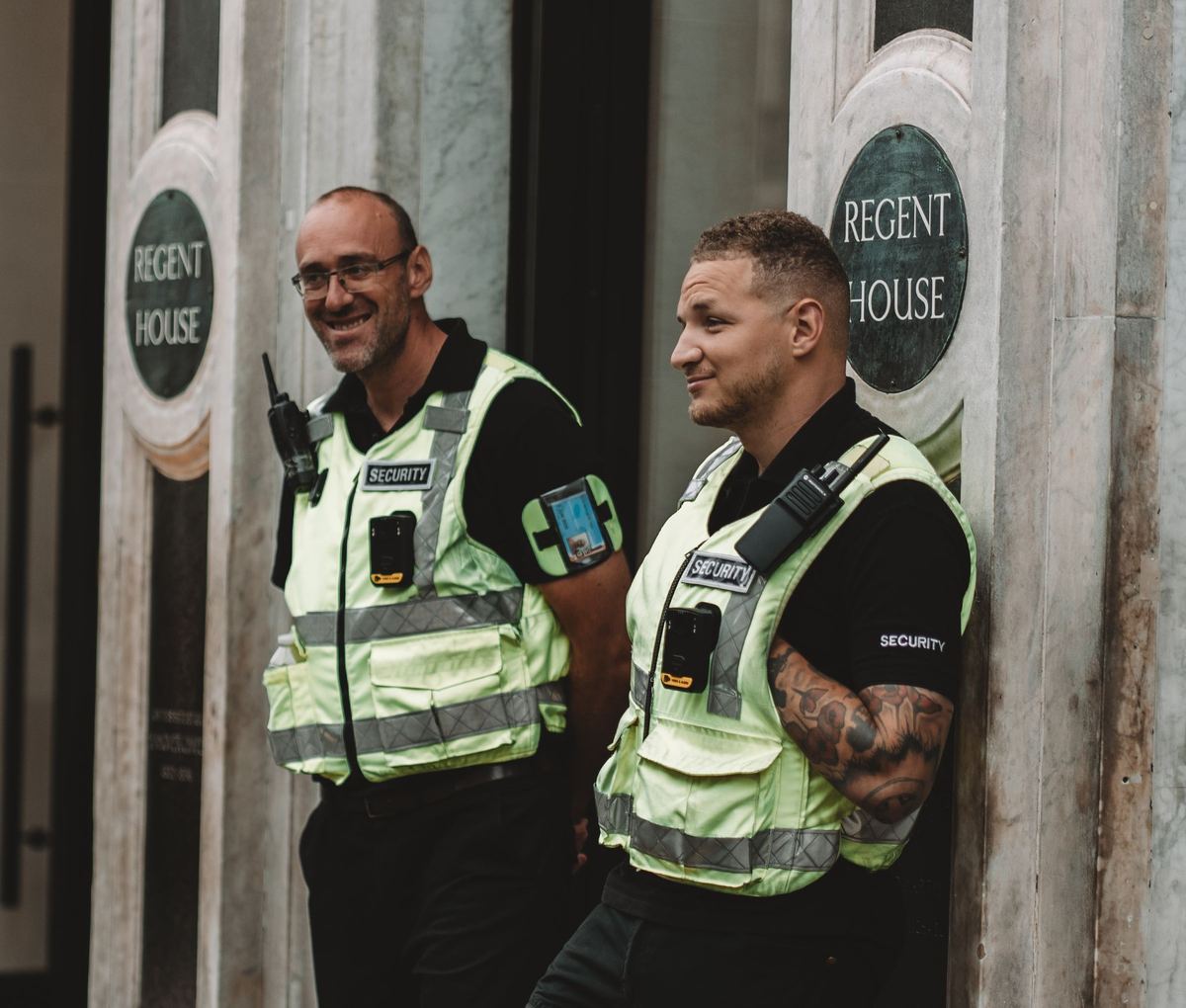 Two security officers in hi-vis vests with radios and body cameras stand at the entrance of an office building in London.