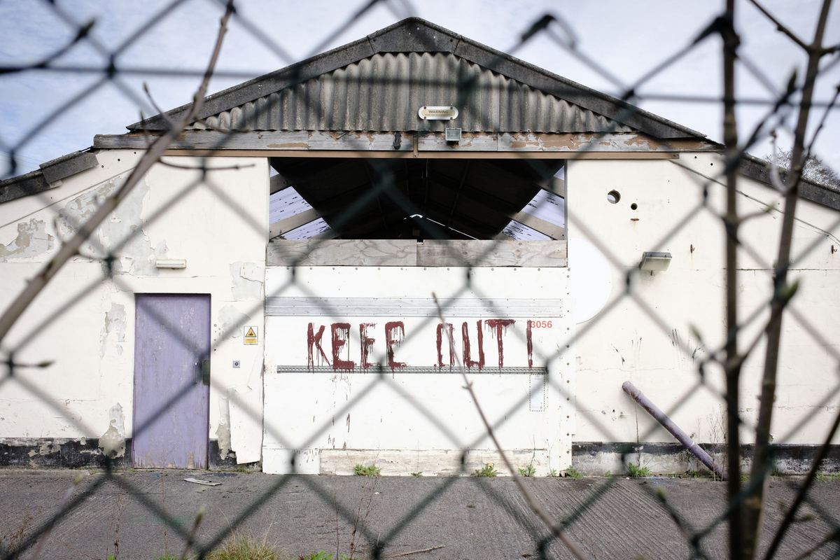 A derelict warehouse in Wotton-under-Edge, England, with KEEP OUT painted on the wall, seen through a wire fence.
